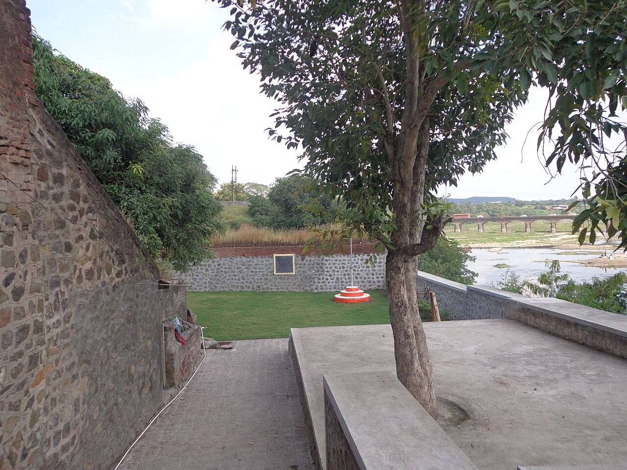 The Rajguru Wada memorial site at Rajgurunagar: stone walls, a lawn with a flag base and a commemorative plaque, overlooking the Bhima river and a bridge