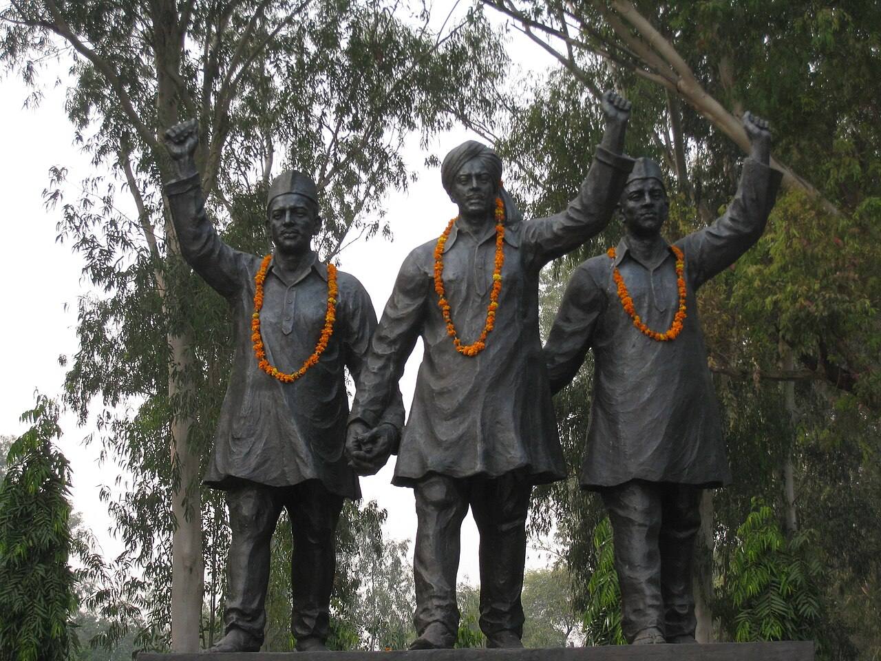 Bronze statues of Bhagat Singh, Rajguru and Sukhdev with raised fists and marigold garlands, standing among trees at a memorial site